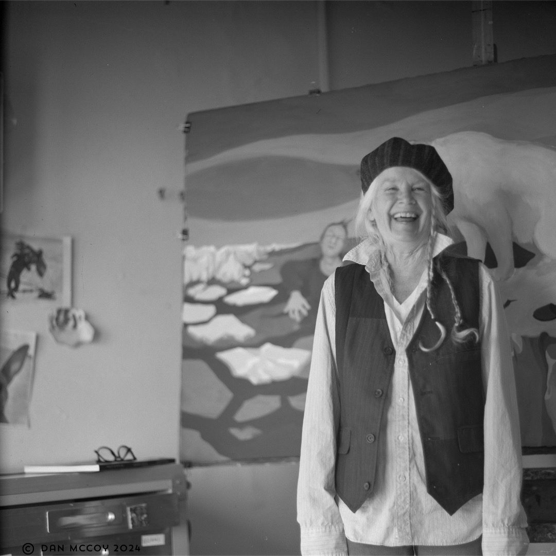 Black-and-white square-format portrait of artist Julie Speed laughing in her Marfa studio, wearing a beret and long braids, standing in front of one of her large paintings.