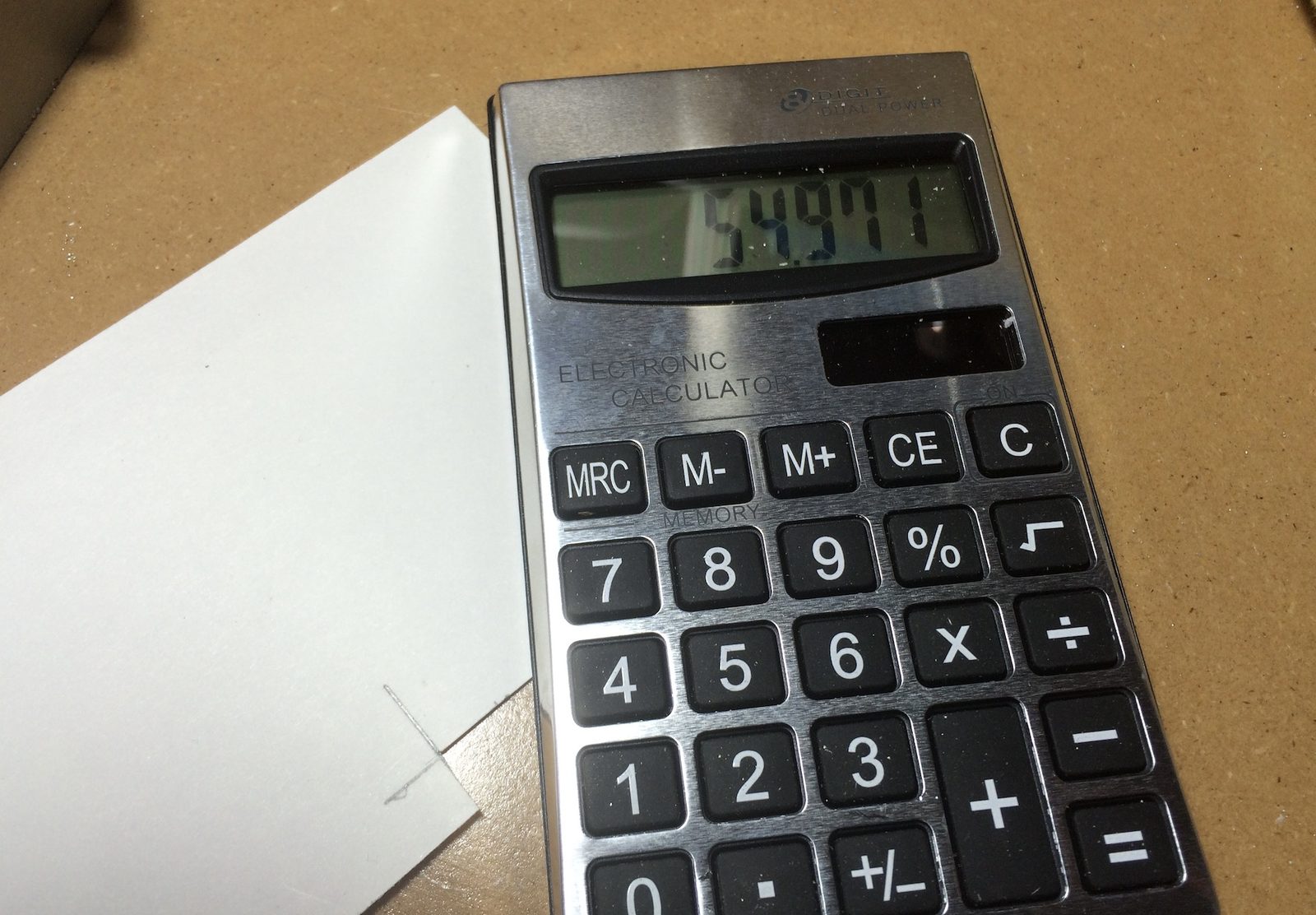 A brushed-metal dual-power electronic calculator sitting on a workbench, its display reading 54.911, next to a sheet of paper with a pencil sketch for a silver ring measurement.