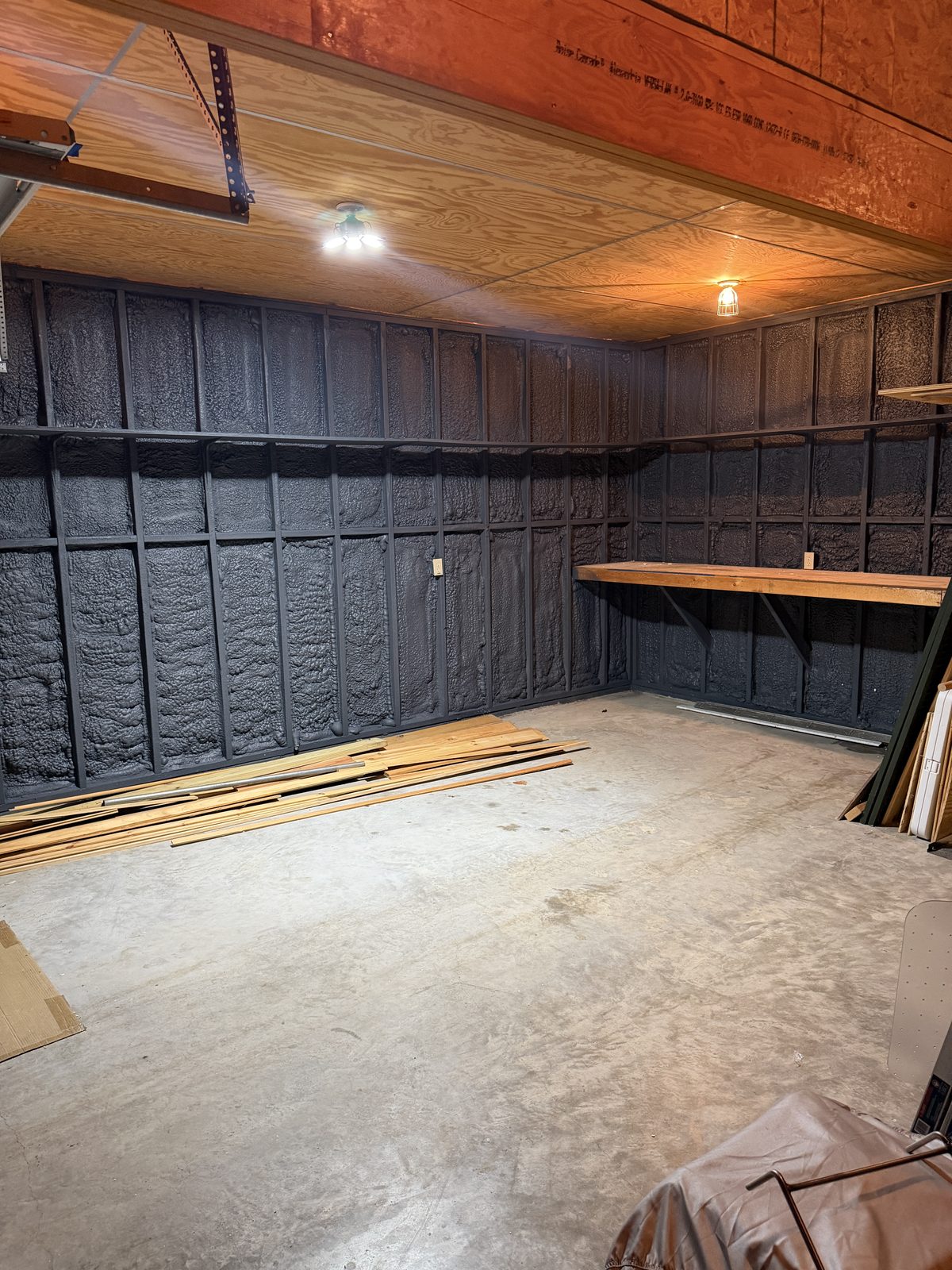 Closed-cell spray foam sprayed between the wall studs of the workshop, cured rough and charcoal-gray, filling every stud bay tight to the framing.