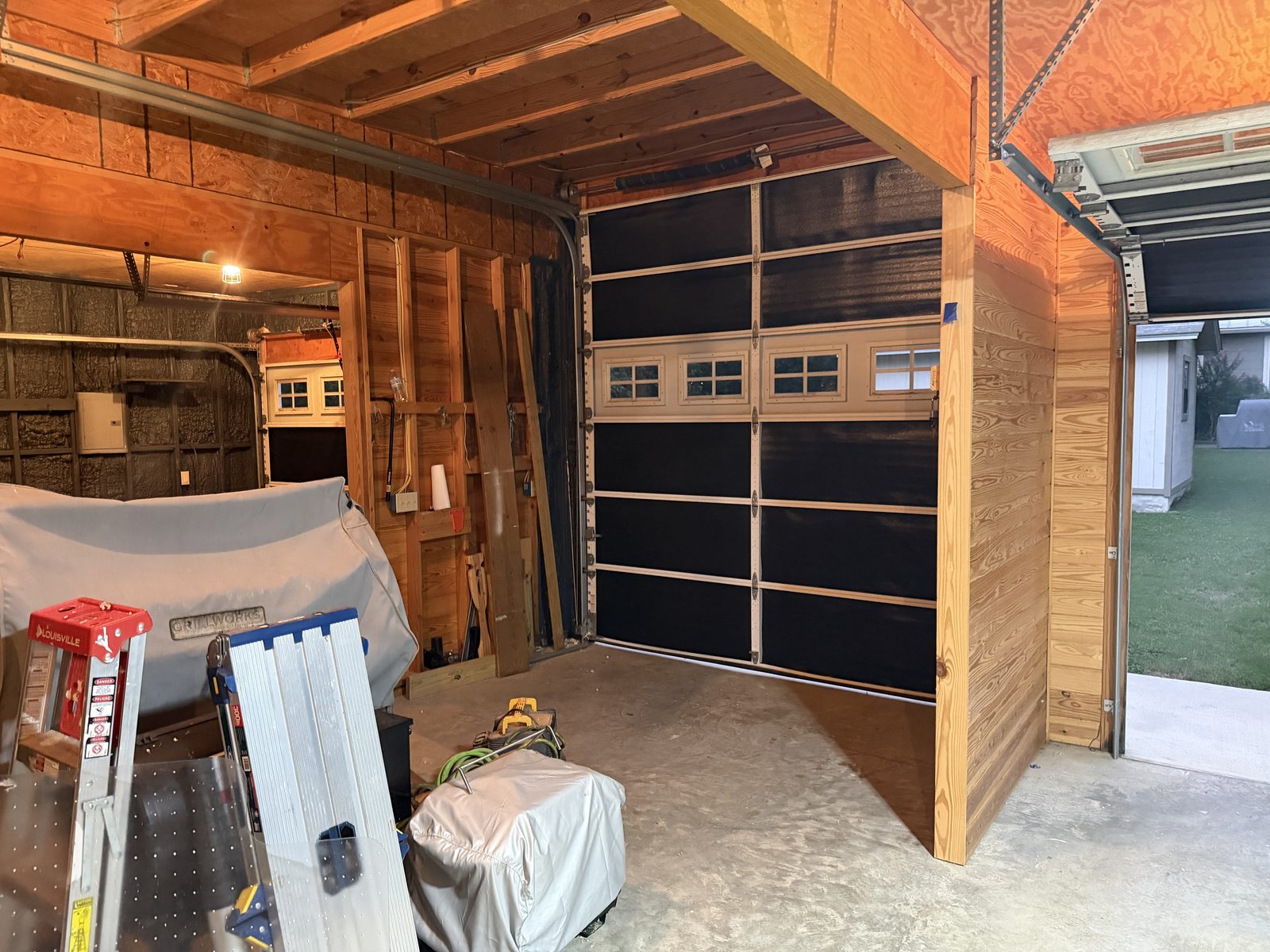 The workshop mid-remodel: bare plywood walls, a black glass-and-panel garage door, concrete floor, ladder and gear staged, the roll-up door open to green grass outside.