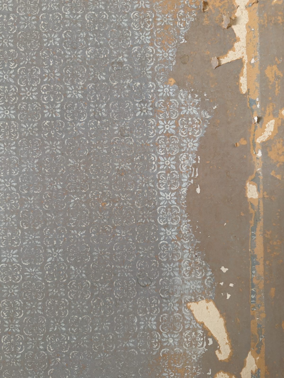 An older stripped-back section of the same wall: a gray-blue ground covered edge to edge in a fine white floral-and-scroll repeat, with ragged tan paper still clinging along the top edge.